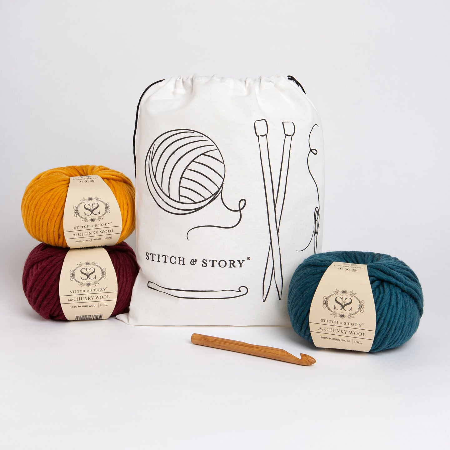 A Stitch & Story Cotton Project bag is placed at the back with 3 balls of 100% merino wool The Chunky Wool, in mustard yellow, sangria red, emerald green and a bamboo crochet hook placed in front of it.