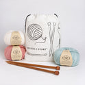 A Stitch & Story Cotton Project bag is placed at the back with 3 balls of 100% merino wool The Chunky Wool, in Pure White, Dust Pink and Iced Mint and a pair of bamboo knitting needles placed in front of it.