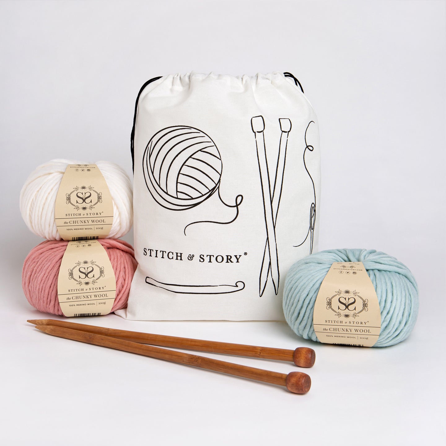 A Stitch & Story Cotton Project bag is placed at the back with 3 balls of 100% merino wool The Chunky Wool, in Pure White, Dust Pink and Iced Mint and a pair of bamboo knitting needles placed in front of it.
