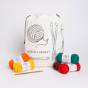 A Stitch & Story Cotton Project bag is placed at the back with 5 balls of Cotton one, in bright red, green, orange, yellow and brown, and a bamboo crochet hook placed in front of it.