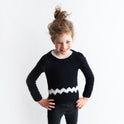 A girl is wearing the Charlie Brown kid sweater, knitted from the knitting kit by Stitch & Story. The long-sleeve sweater is in Black with a graphic White chevron detail across the bottom of the sweater.