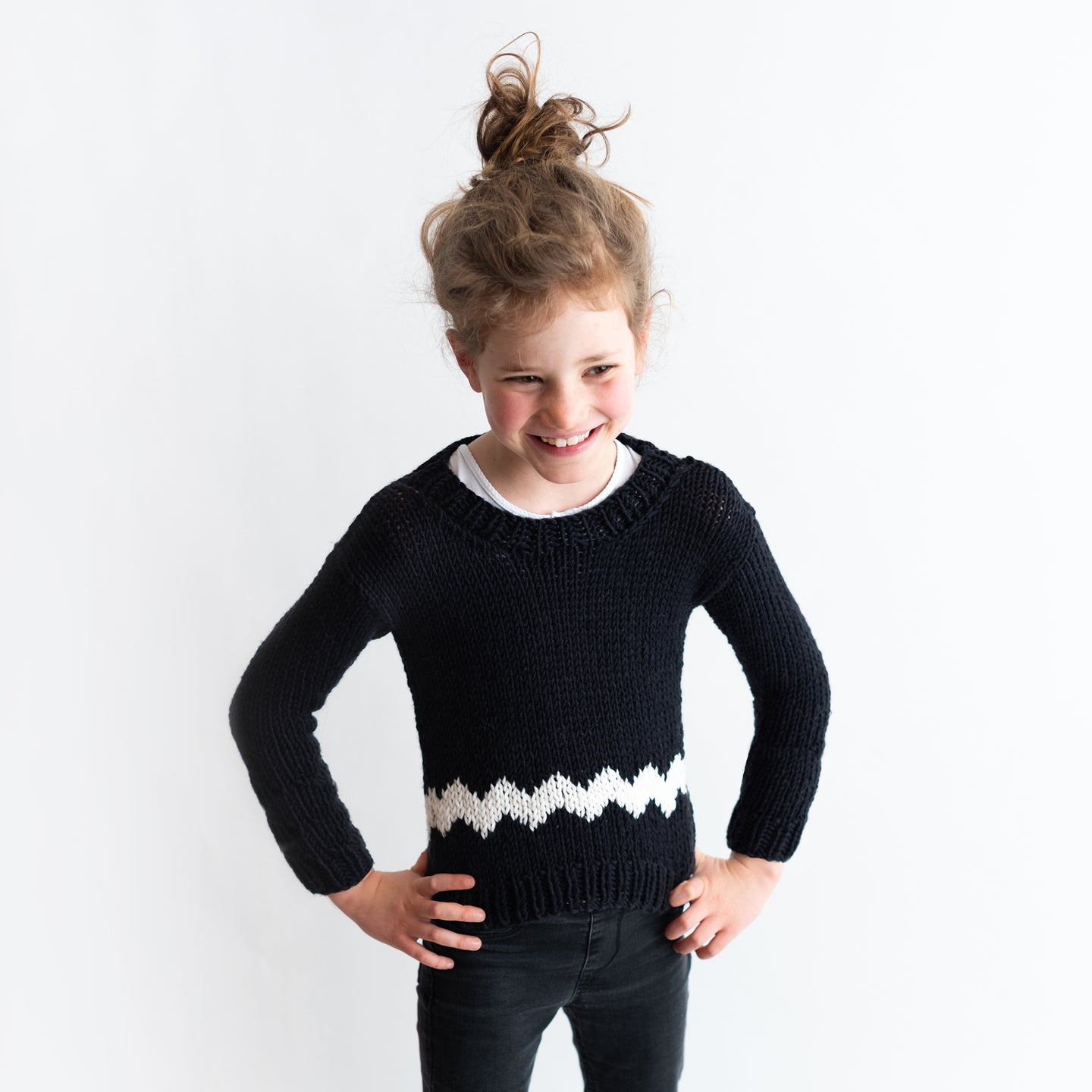 A girl is wearing the Charlie Brown kid sweater, knitted from the knitting kit by Stitch & Story. The long-sleeve sweater is in Black with a graphic White chevron detail across the bottom of the sweater.