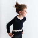 A laughing girl is wearing the Charlie Brown kid sweater, knitted from the knitting kit by Stitch & Story. The long-sleeve sweater is in Black with a graphic White chevron detail across the bottom of the sweater.