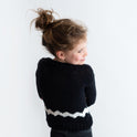 It shows the back of a girl wearing the Charlie Brown kid sweater, knitted from the knitting kit by Stitch & Story. The long-sleeve sweater is in Black with a graphic White chevron detail across the bottom of the sweater.