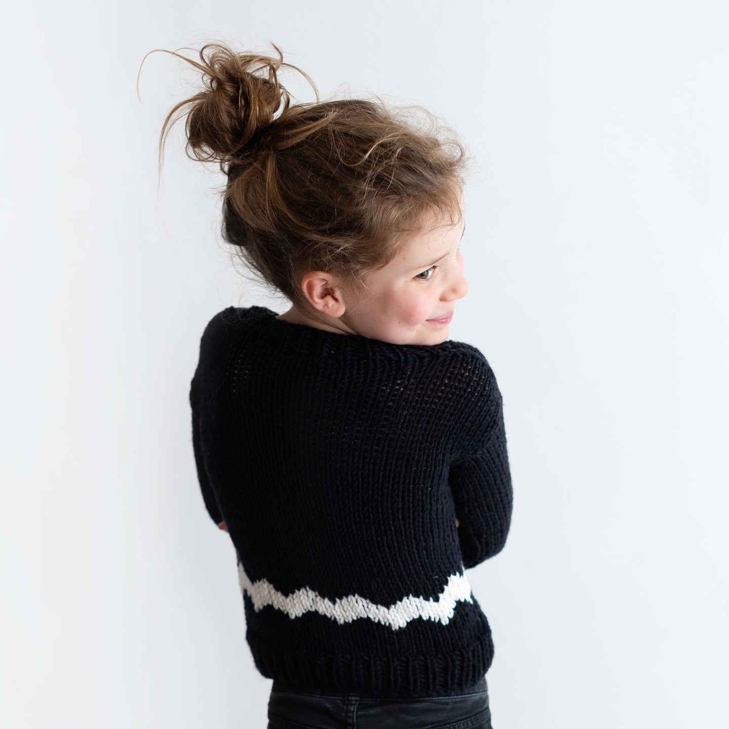 It shows the back of a girl wearing the Charlie Brown kid sweater, knitted from the knitting kit by Stitch & Story. The long-sleeve sweater is in Black with a graphic White chevron detail across the bottom of the sweater.