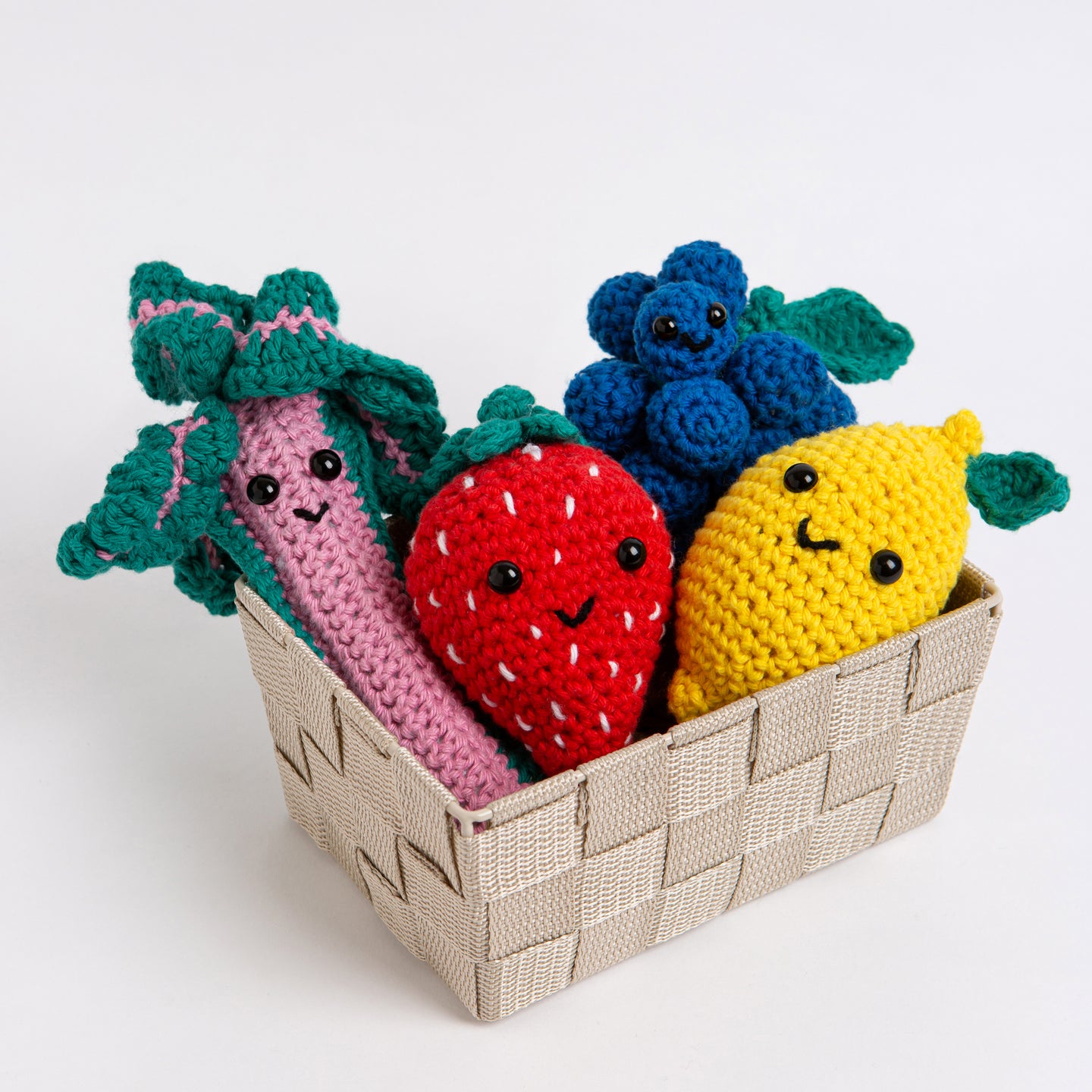 4 Fruit crochet amigurumi toys are all placed in a small basket with their smiling faces facing outwards. They include Aubrey the Strawberry in Red Apple Cotton One, Lemmy the Lemon in Daisy Yellow in Cotton One, Bard the Rhubarb in Rose Pink in Cotton One and Merlot the Grape in Royal Blue in Cotton one.