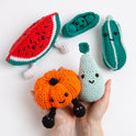 A hand is holding up 2 crochet amigurumi fruit toys, and all remaining 3 are laid out on the table under the hand. All are smiling at the camera in a super colourful photo