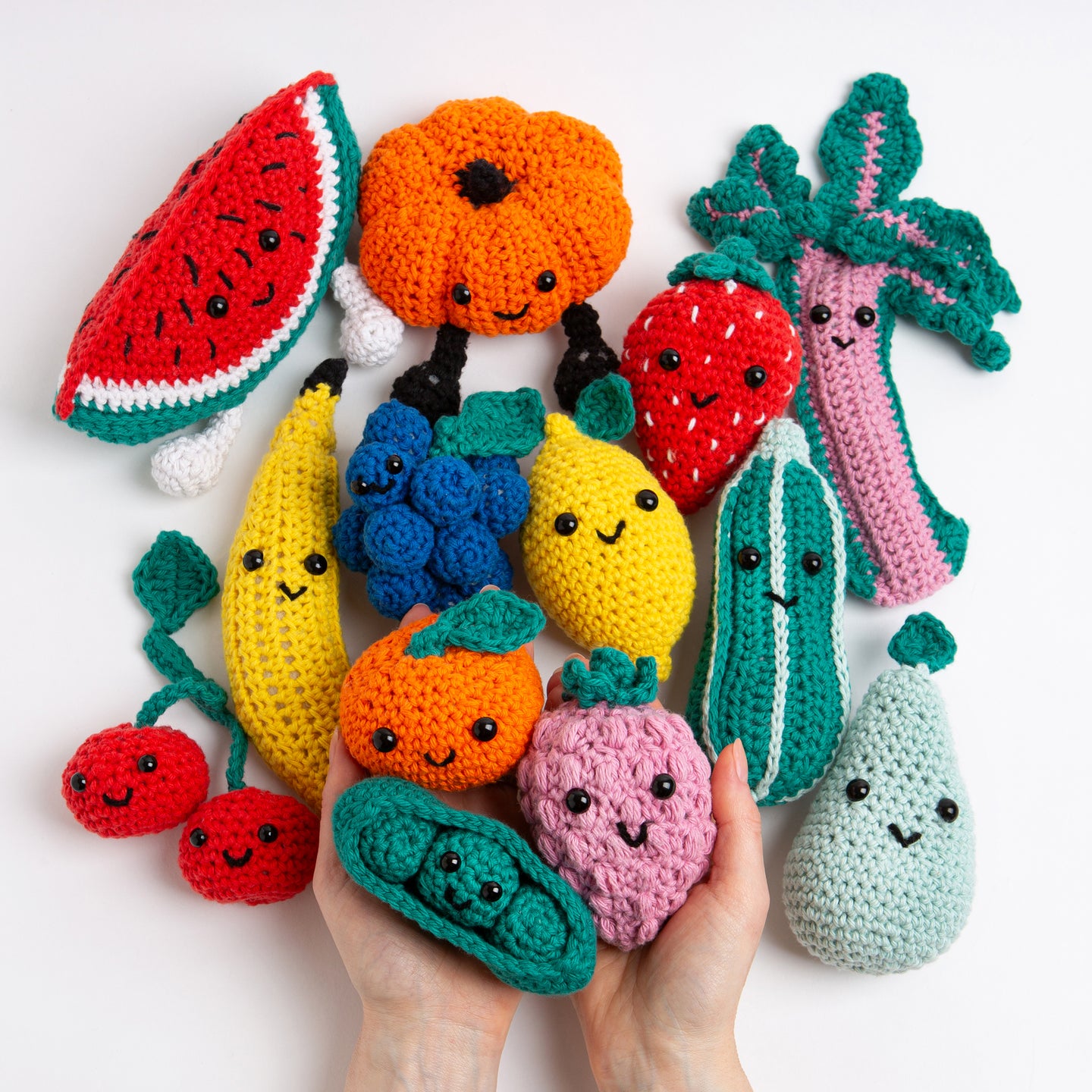 A hand is holding up 3 crochet amigurumi fruit toys, and all remaining 10 are laid out on the table under the hand. All are smiling at the camera in a super colourful photo