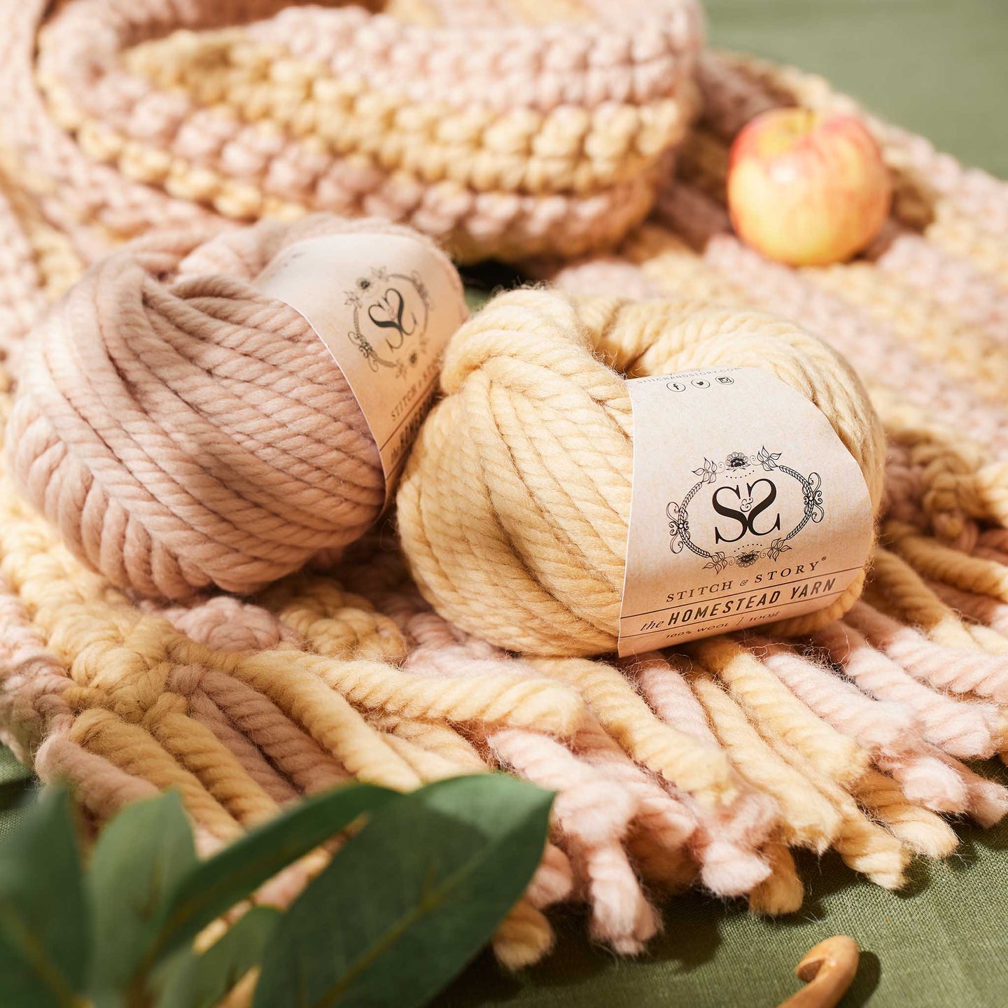 2 balls of 100% Super Chunky wool Homstead Yarn from Stitch & Story, in Sand Dune and Pink Peony are photographed with a piece of crochet fabric on a green linen fabric, giving a sense of back to nature.