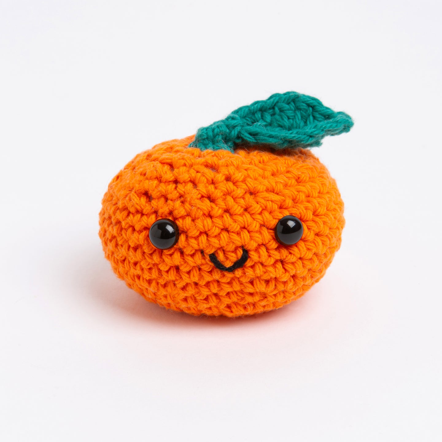 An orange toy made with Stitch & Story Cotton One in Deep Orange has a joyful smiley faces, with a Forest Green leaf at the top, on a white background