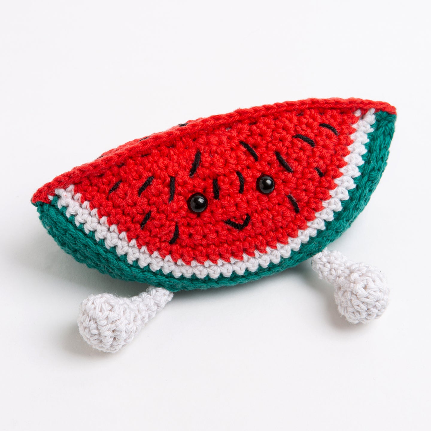 A watermelon toy made with Stitch & Story Cotton One in Red Apple and Forest Green has a joyful smiley face, and a pair of dangly feet in Paper White, on a white background