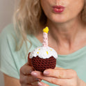 Someone is holding a crochet candle lit sugar glazed cupcake and blowing on the candle, after making a wish.