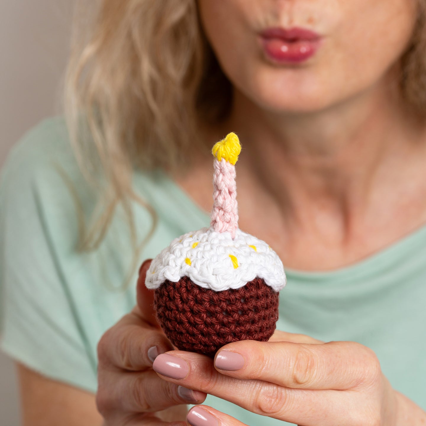 Someone is holding a crochet candle lit sugar glazed cupcake and blowing on the candle, after making a wish.