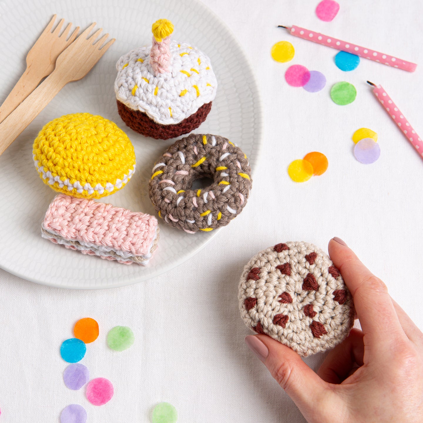 A hand is taking away a crochet chocolate chip cookie, from a plate with a sprinkle donut, pink wafer biscuit, yellow lemon macaroon and a candle-lit sugar glazed cupcake. All crocheted and ready for celebration.