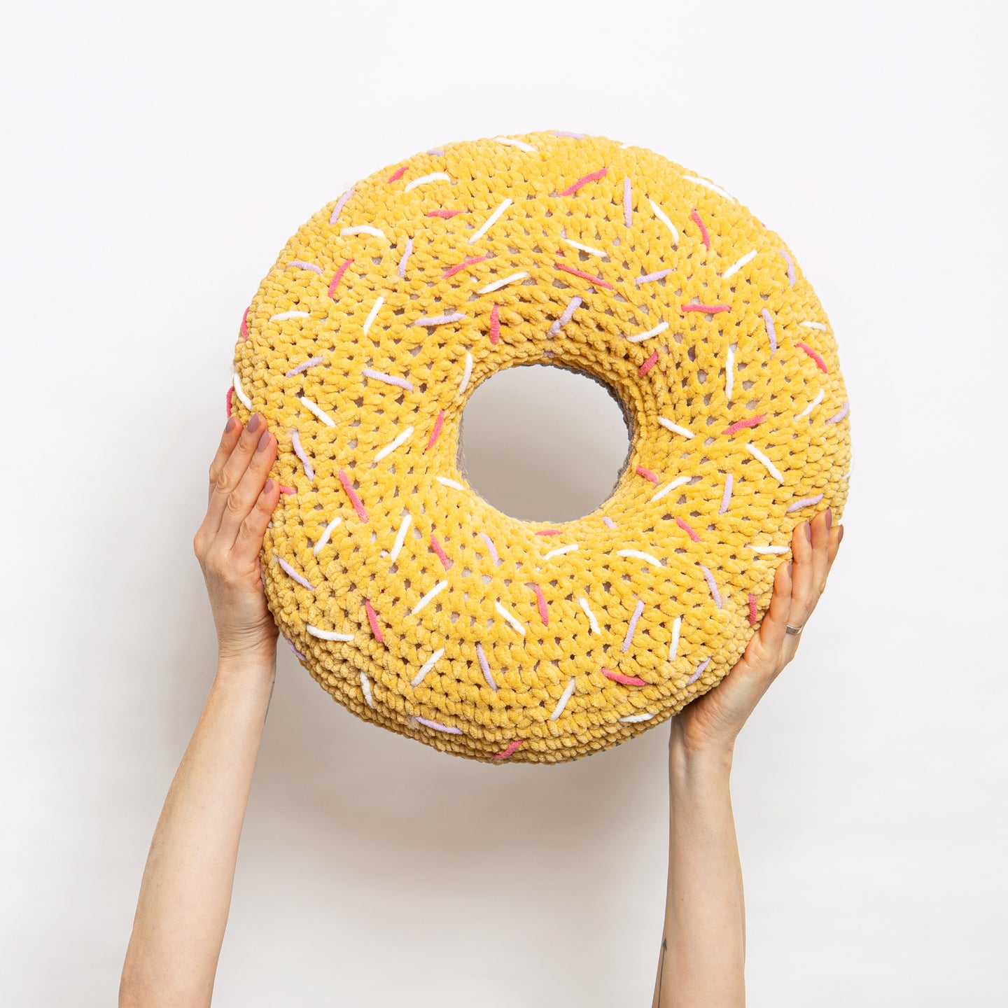 Someone is holding high up a giant round ring yellow plush cushion with pink, lilac and white sprinkles, all made with Stitch & Story Roly Poly Plush. 