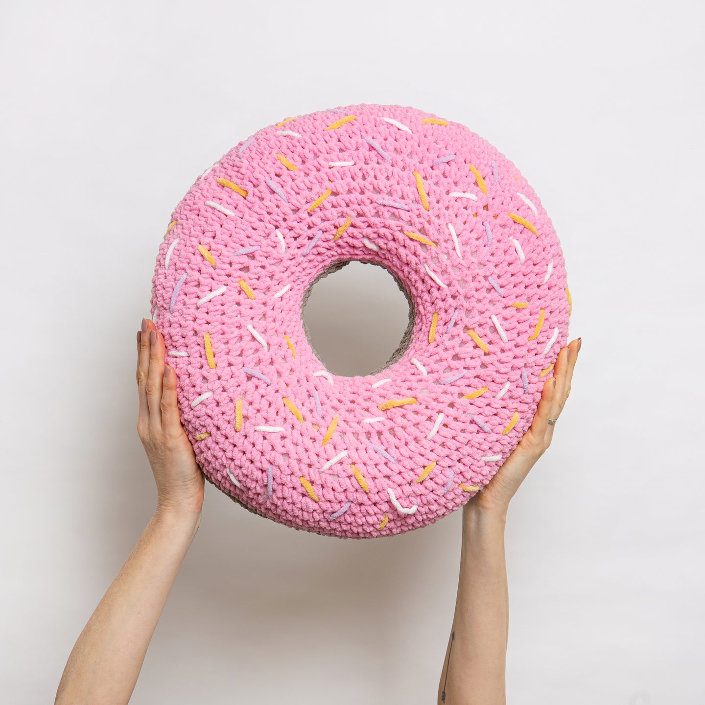 Someone is holding high up a giant round ring pink plush cushion with yellow, lilac and white sprinkles, all made with Stitch & Story Roly Poly Plush. 