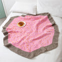 a pink centred doughnut blanket is placed stylishly on a bed with 2 white pillows. A plate with a cinnomon roll is placed on top of the blanket as this set up is ready for a cosy night in.