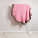 A draped pink doughnut blanket is styled on a white railing, against a grey wall. The blanket is round and softly draped over.
