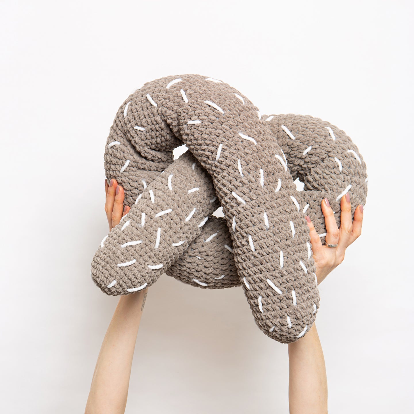 Someone is holding high up a giant light grey twisted pretzel plushie cushion with white sprinkles, all made with Stitch & Story Roly Poly Plush. 