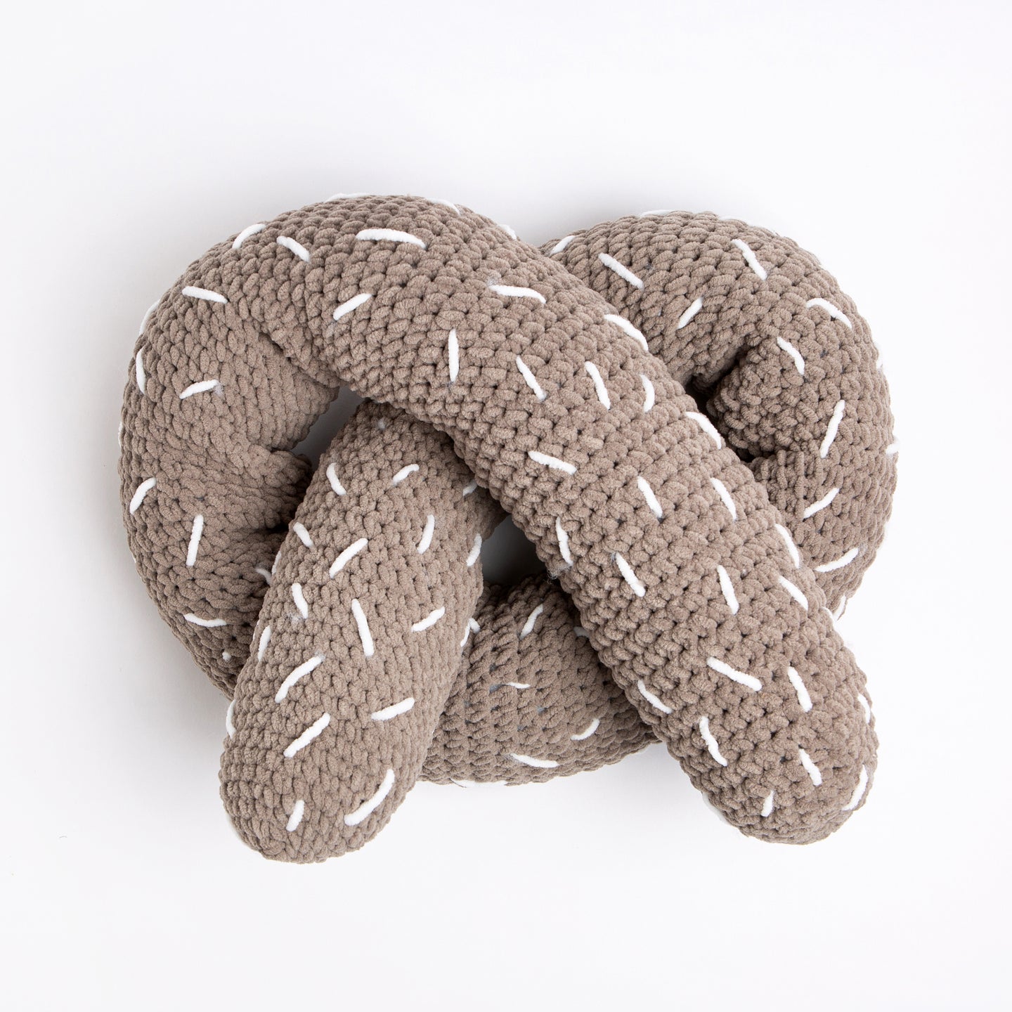 A giant light grey plush pretzel cushion with white sprinkles, all made with Stitch & Story Roly Poly Plush is placed on a white background. 