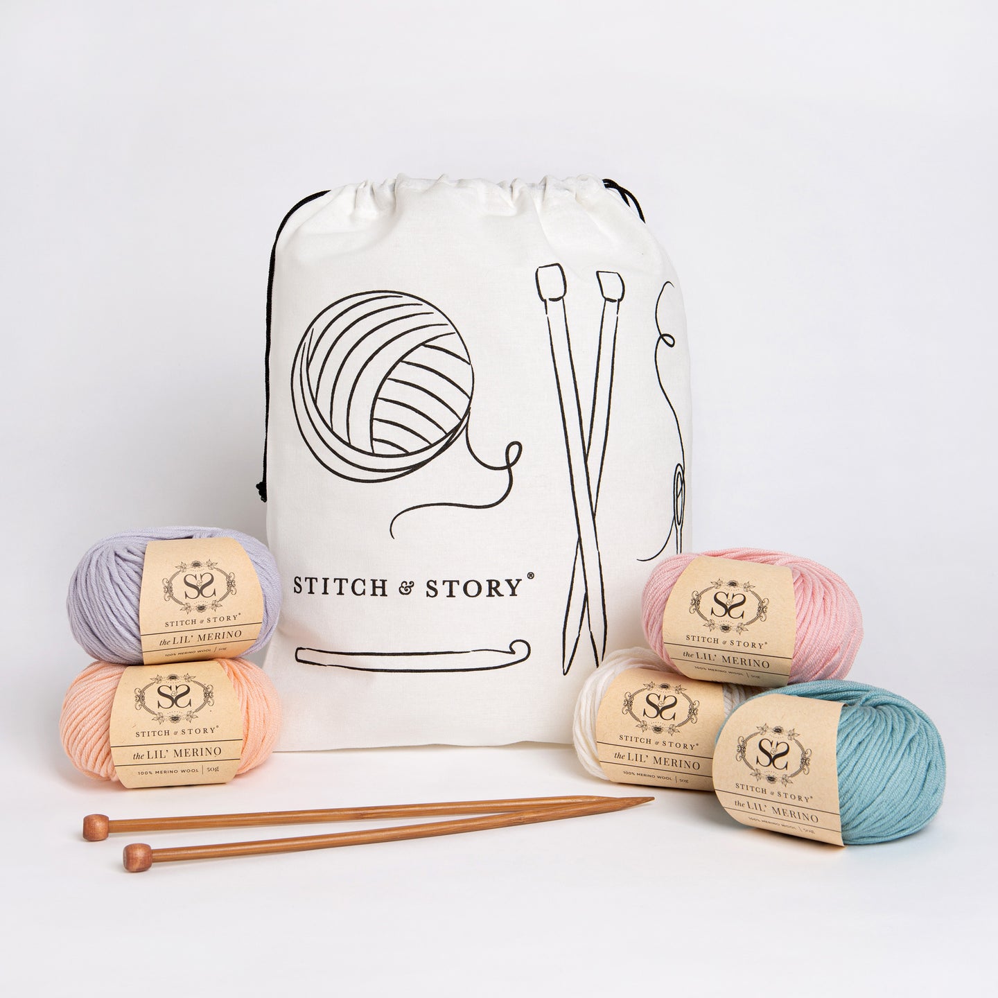 A Stitch & Story Cotton Project bag is placed at the back with 5 balls of Lil' Merino, in Dove Grey, Peach Pink, Doe Pink, Bright White and Soft Teal, and a pair of bamboo knitting needles placed in front of it.