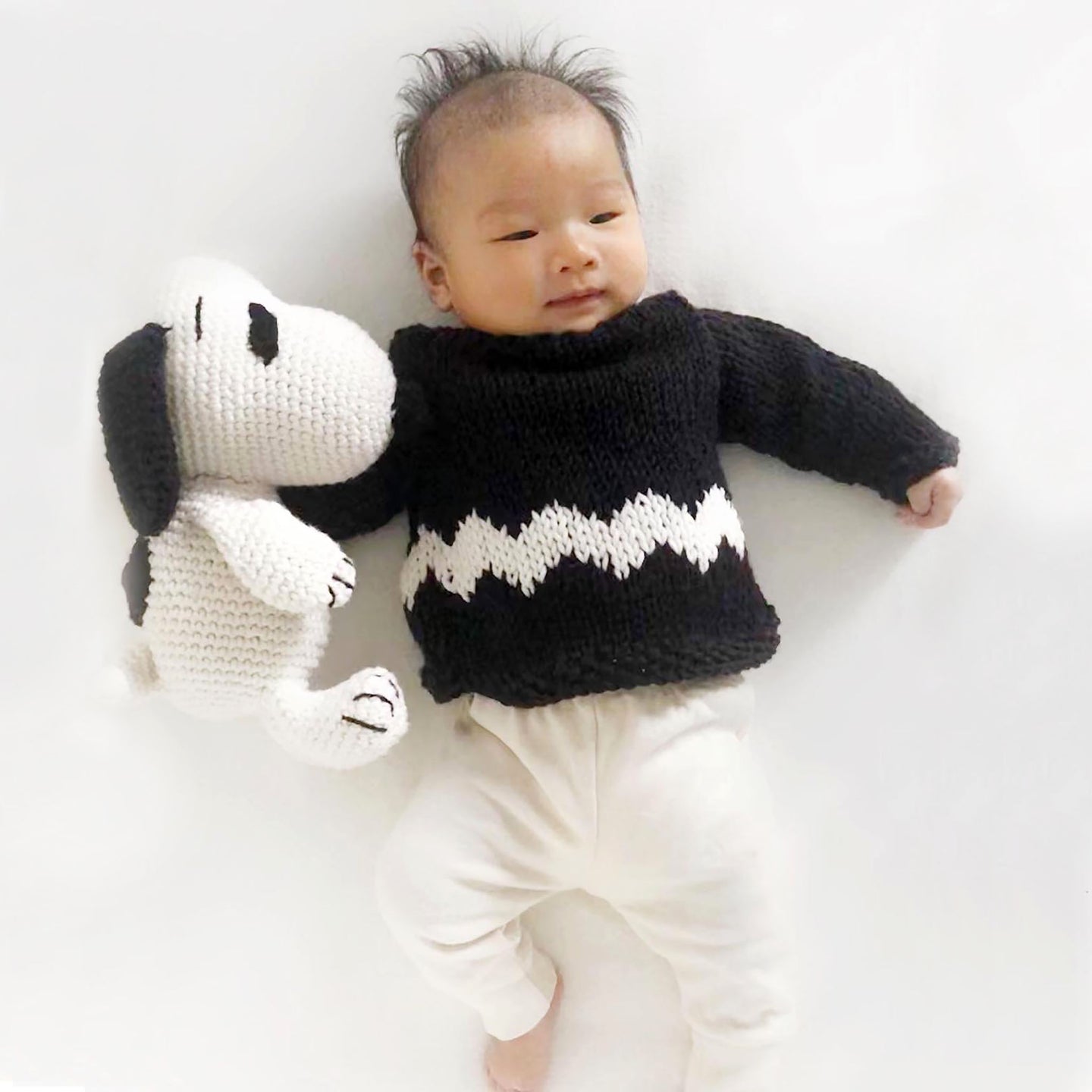 A baby is lying down, wearing a black jumper with a white graphic chevron pattern on it, the Charlie Brown baby's sweater knitting kit from Stitch & Story. Next to the baby is also the Snoopy crochet toy, another available kit from the same collection.