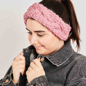 A woman is looking cosy wearing her Dust Pink Chunky Cable Headband, knitted from Stitch & Story's knitting kit using 100% super chunky merino wool.