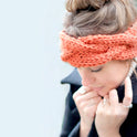 A woman is looking cosy wearing her orange Chunky Cable Headband, knitted from Stitch & Story's knitting kit using 100% super chunky merino wool.