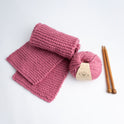 A folded Grazier Scarf in Dust Pink, knitted from Stitch & Story's Grazier scarf beginner knitting kit, laid out next to a ball of 100% Merino The Chunky Wool and a pair of 12mm bamboo knitting needles.