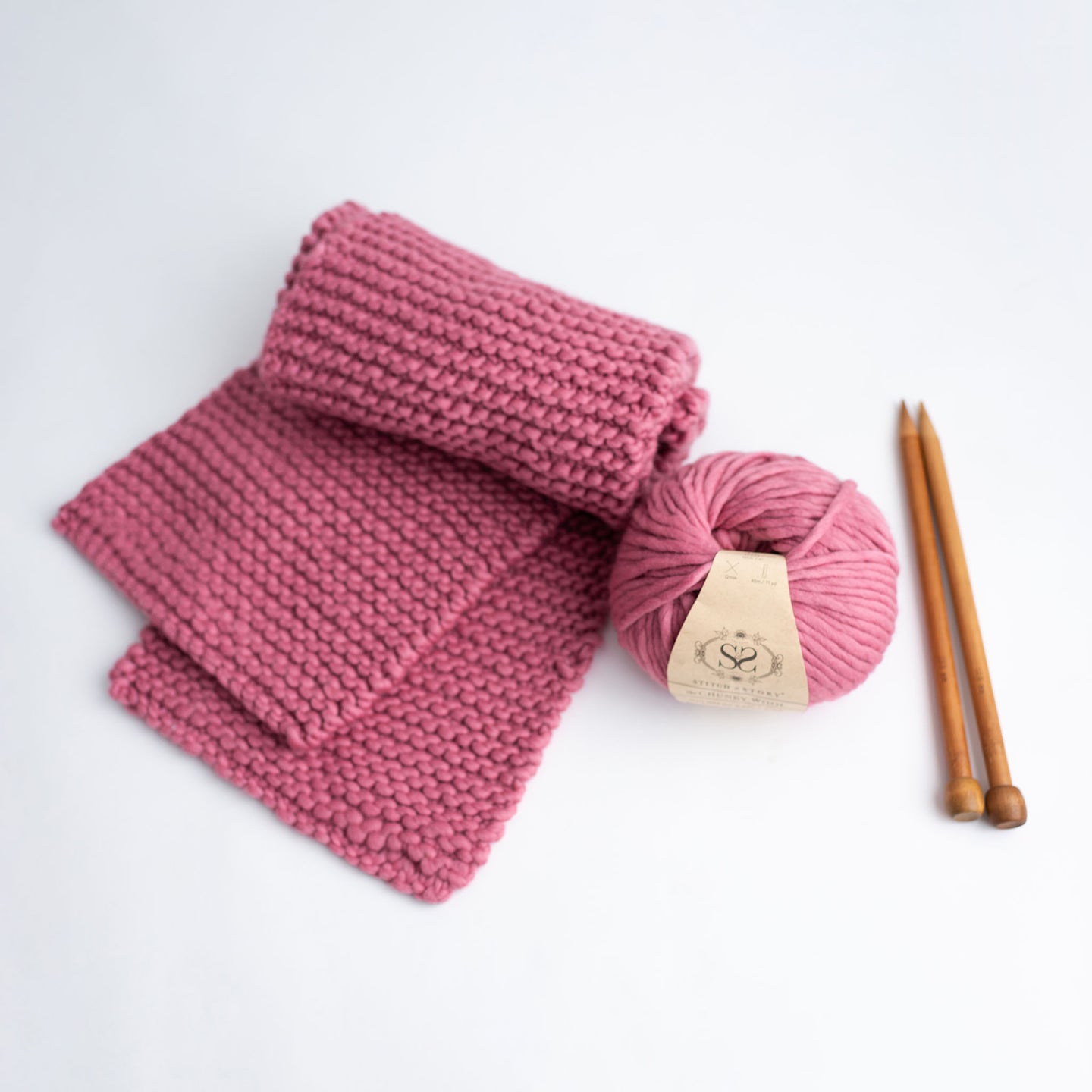 A folded Grazier Scarf in Dust Pink, knitted from Stitch & Story's Grazier scarf beginner knitting kit, laid out next to a ball of 100% Merino The Chunky Wool and a pair of 12mm bamboo knitting needles.