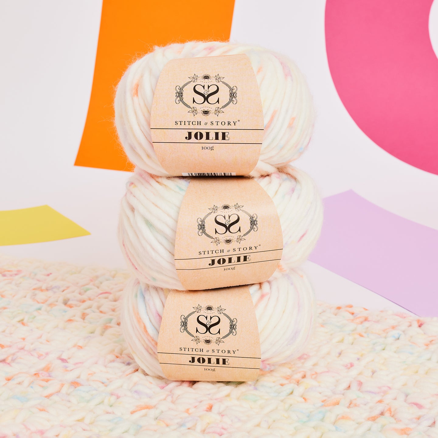 3 balls of Jolie Yarn in Pastel Rainbow is stacked on each other, placed on top of a piece of crochet fabric in the super chunky Jolie Yarn, in front of a vibrant background