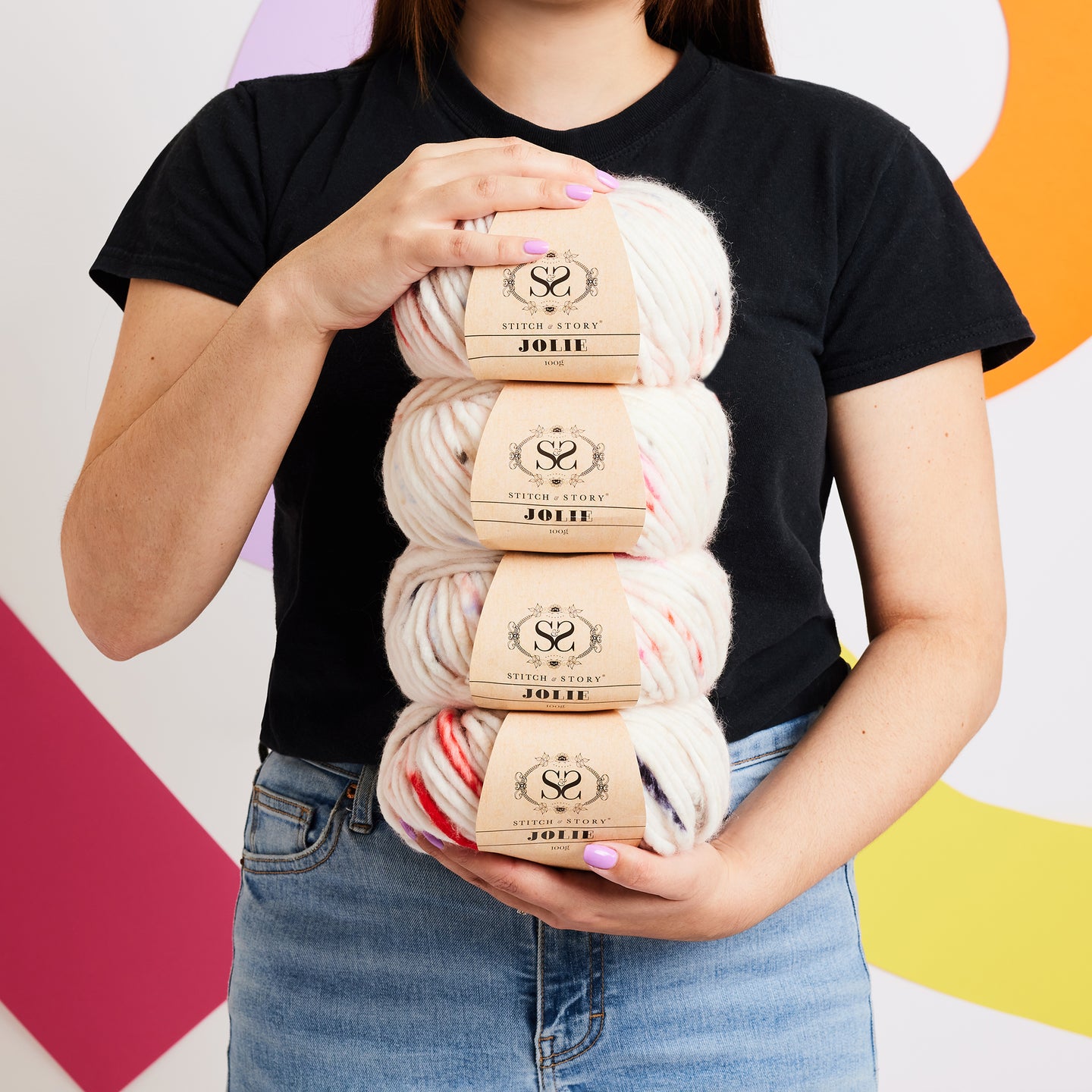 A person in a black tshirt is holding 4 balls of Jolie Yarn in Wild Flowers in their hand showing you how chunky this yarn is and how great it is for beginner knitters, stood in front of a vibrant background.