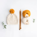 A Luca Pom hat knitted in Ivory white with a mustard yellow pom pom, laid next to a pair of 12mm bamboo knitting needles and 2 balls of Stitch & Story Chunky Wool Yarn.
