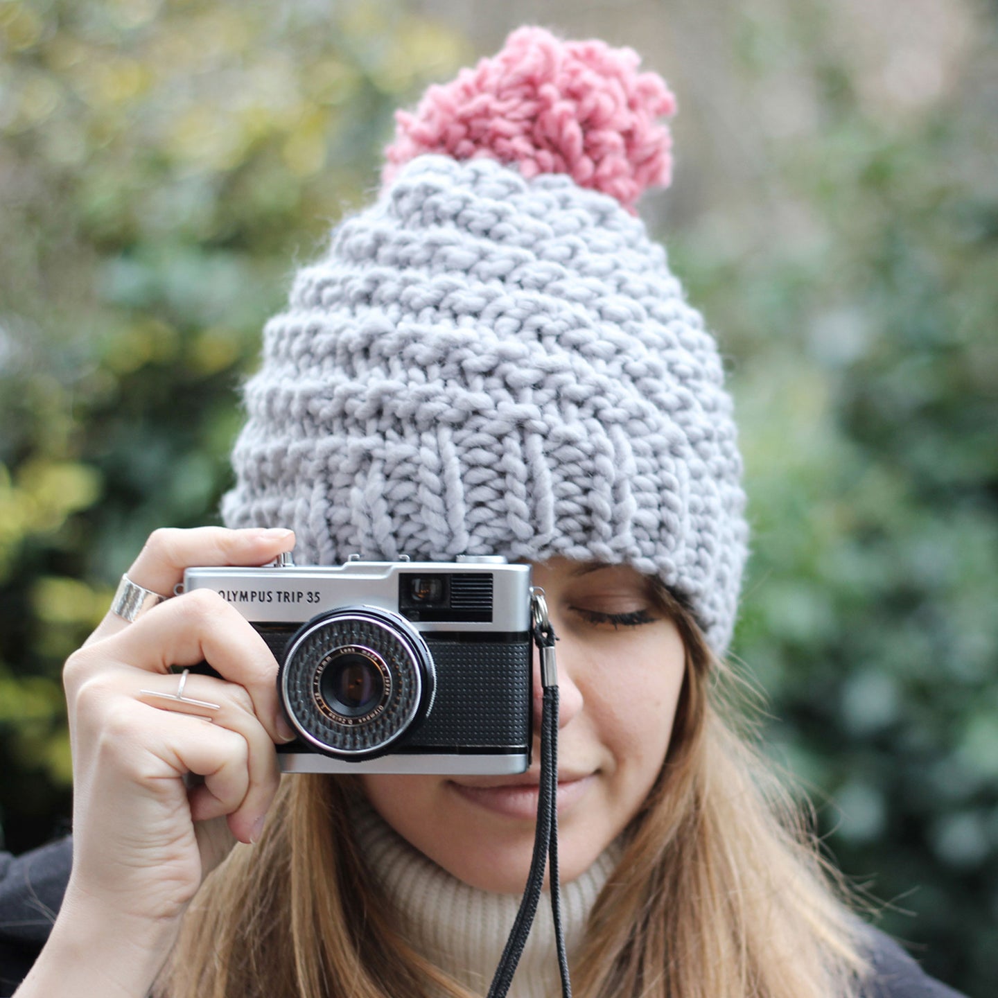 A woman is wearing the Luca Pom Hat in Stormy Grey with a Pink pom pom, knitting from a Stitch & Story kit, with a camera held to her face.