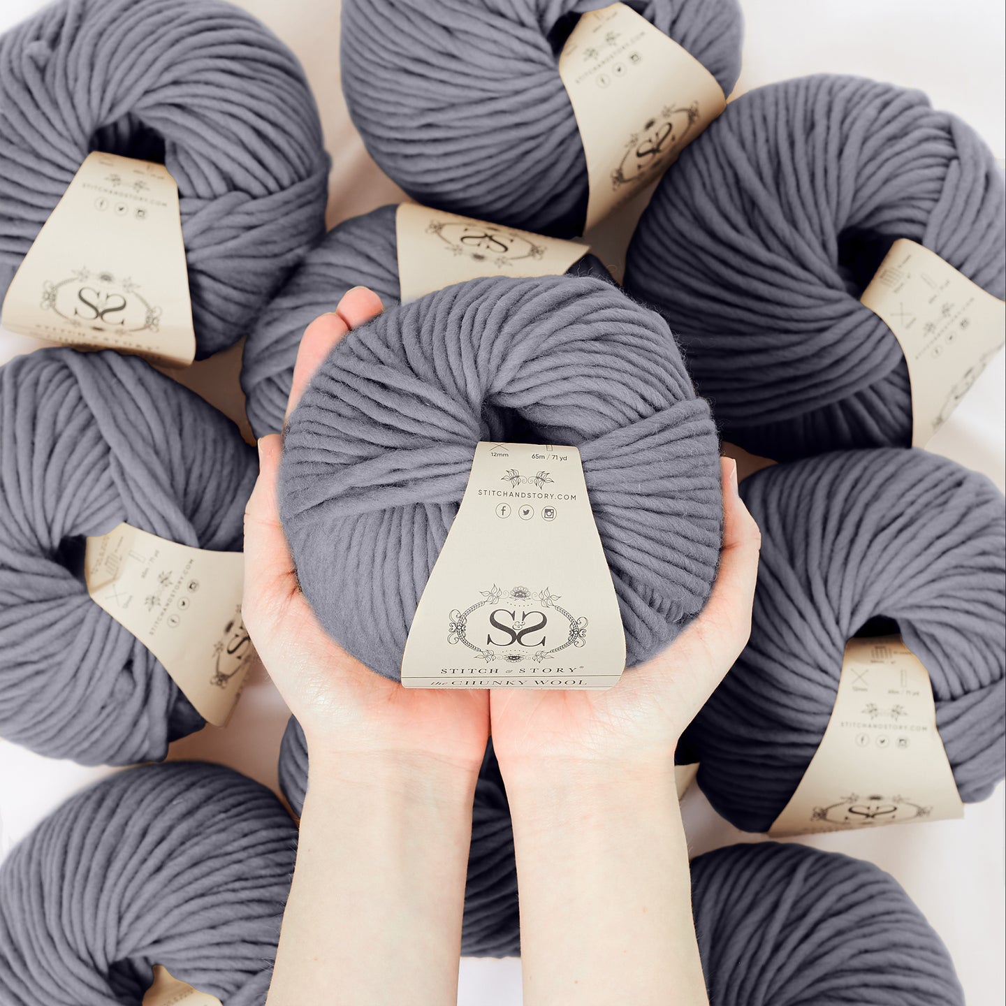 A hand is holding a ball of 100% merino wool Stitch & story The Chunky Wool in front of the camera, above another 9 balls of the same yarn. The colour shown here is fossil grey