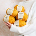 a person is holding 5 balls of Mustard Yellow 100% merino wool The Chunky Wool from Stitch & story in a cosy manner, ready for a day of knitting.