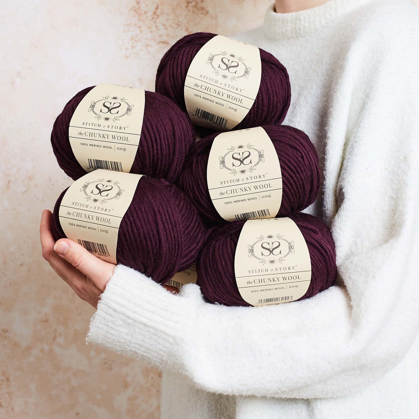 a person is holding 5 balls of Rich plum 100% merino wool The Chunky Wool from Stitch & story in a cosy manner, ready for a day of knitting.