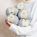 a person is holding 5 balls of Stormy Grey 100% merino wool The Chunky Wool from Stitch & story in a cosy manner, ready for a day of knitting.