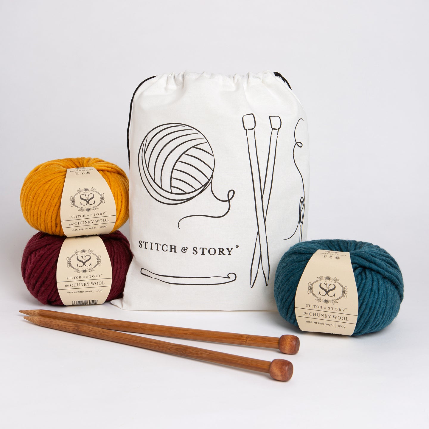 A Stitch & Story Cotton Project bag is placed at the back with 3 balls of 100% merino wool The Chunky Wool, in Mustard Yellow, Sangria Red and Emerald Green and a pair of bamboo knitting needles placed in front of it.