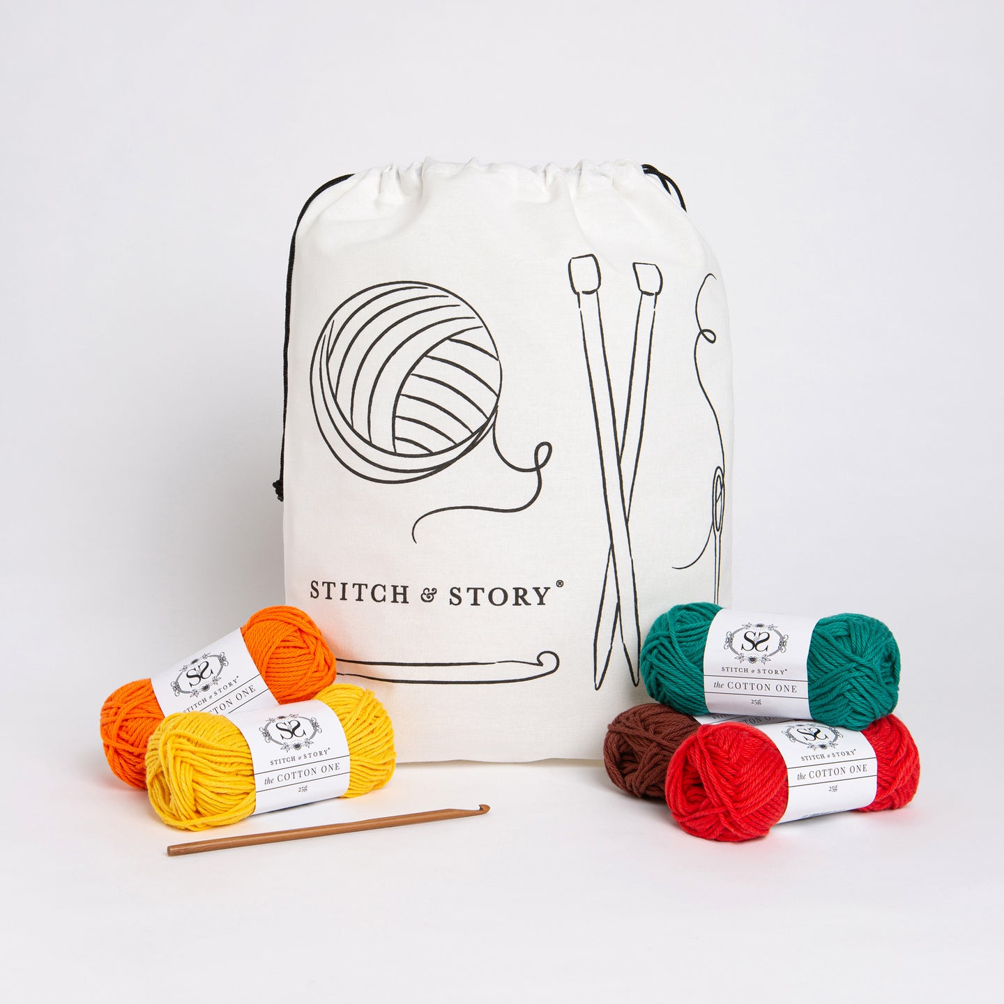 A Stitch & Story Cotton Project bag is placed at the back with 5 balls of Cotton one, in bright red, green, orange, yellow and brown, and a bamboo crochet hook placed in front of it.