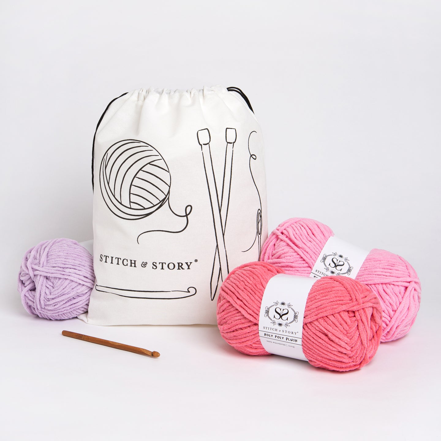 A Stitch & Story Cotton Project bag is placed at the back with 3 balls of roly Poly Plush, in lilac, light pink and dark pink, and a bamboo crochet hook placed in front of it.