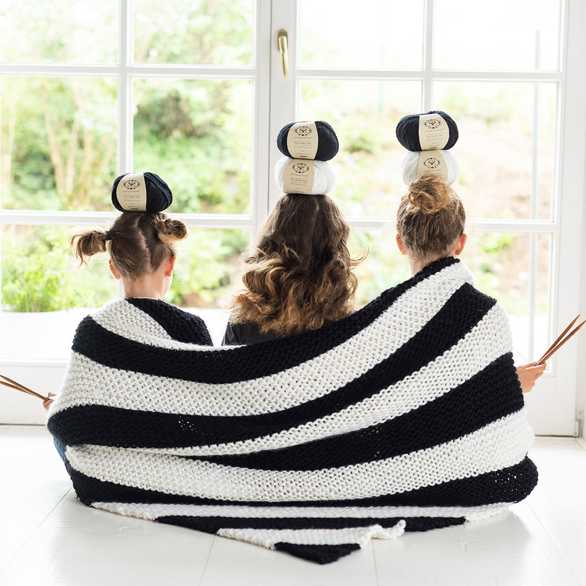 3 girls are playfully balancing balls of Stitch & Story 100% merino wool The Chunky Wool whilst wrapped together in the Snoopy black and white chunky Throw, knitted from the Snoopy Throw knitting Kit.