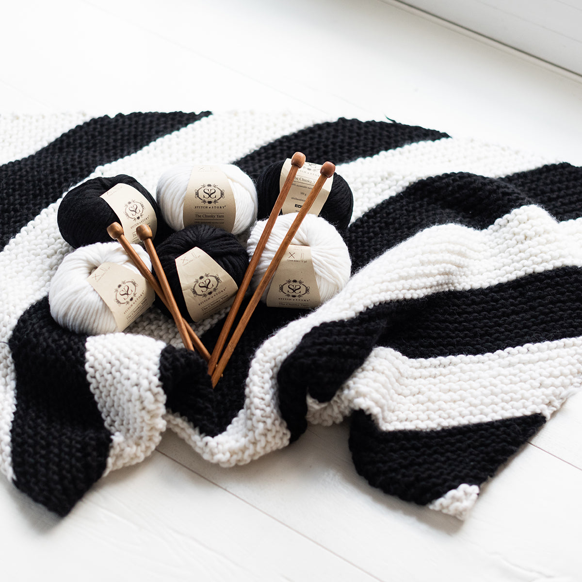 A close up of the Snoopy black and white throw, with 6 balls of The Chunky Wool from Stitch & Story are laid on top of it.