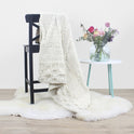 An Ivory White Bounty Throw knitted from 100% super chunky Merino wool is draped over a black chair, next to a table with a vase of flower on it. This is from a beginner friendly Stitch & Story Knitting Kit