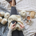 A person is seated with biscuit and tea, a balls of super soft Merino wool The chunky Wool, whilst knitting the Chunky Tassel Throw in Ivory White on a pair of bamboo knitting needles.