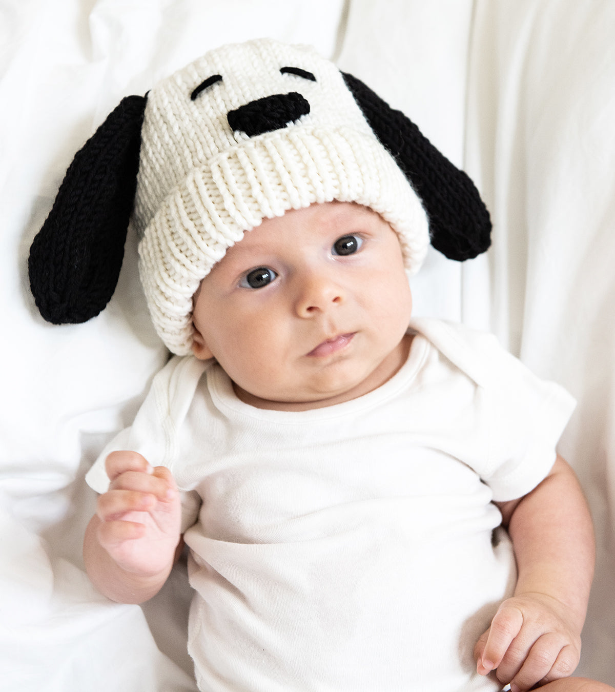 A focused baby is wearing the Snoopy baby hat, made with Stitch & Story's 100% merino wool in white, with black ears.