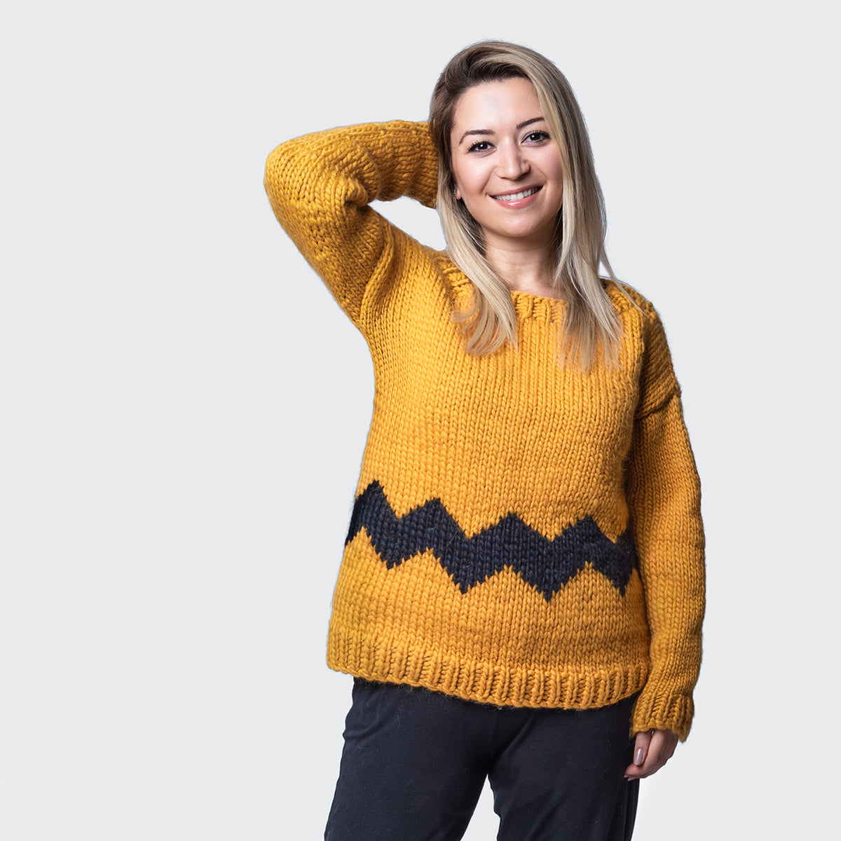 a woman is wearing a cosy fitted Charlie Brown sweater knitted from 100% merino wool The Chunky Wool from Stitch & Story. The sweater is in Mustard yellow with a graphic black chevron pattern at the bottom of the sweater.