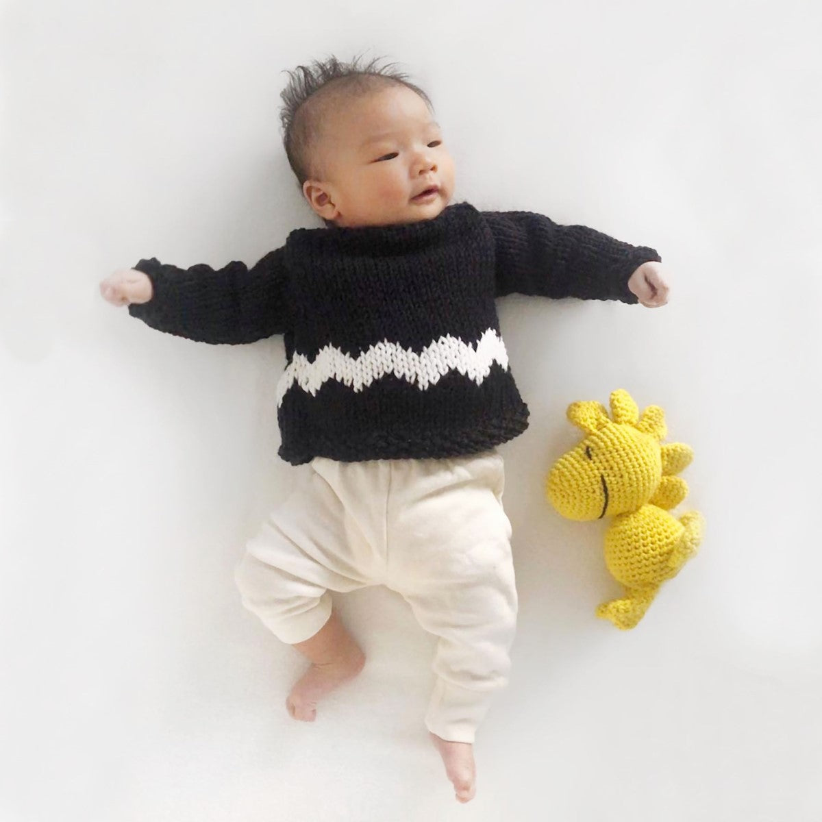 A baby is lying down, wearing a black jumper with a white graphic chevron pattern on it, the Charlie Brown baby's sweater knitting kit from Stitch & Story. Next to the baby is also the Woodstock crochet toy, another available kit from the same collection.