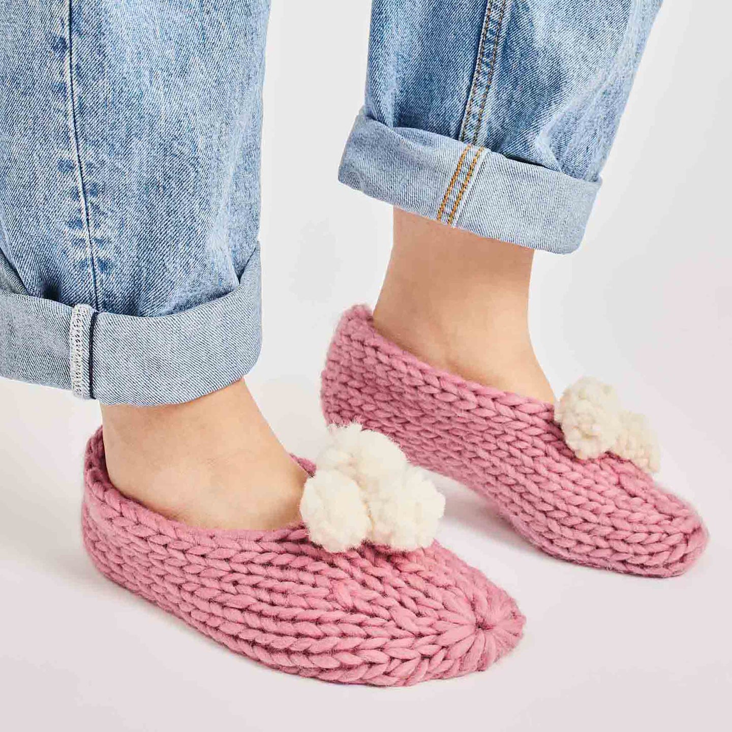 A woman is standing wearing pink lola pom slippers with white pom pom knitted from Stitch & Story beginner knitting kit