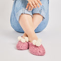 A woman is seated with leg crossed, wearing pink lola pom slippers with white pom pom knitted from Stitch & Story beginner knitting kit